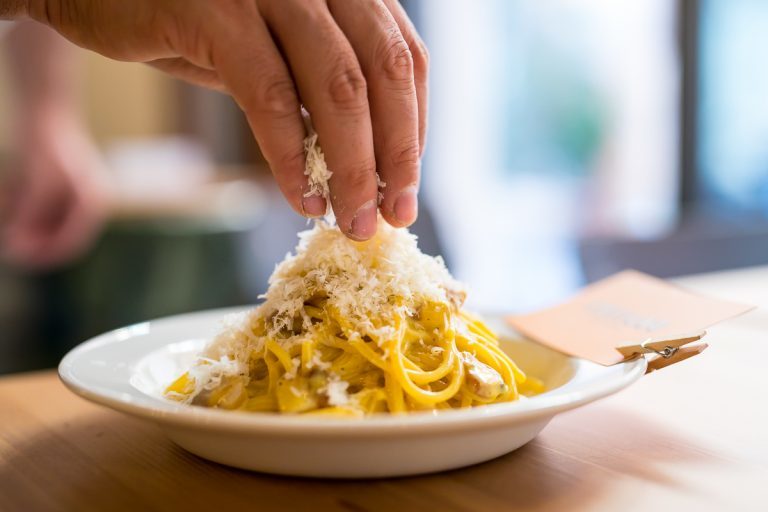 Mano cosparge di formaggio grattugiato su un piatto di pasta alla carbonara